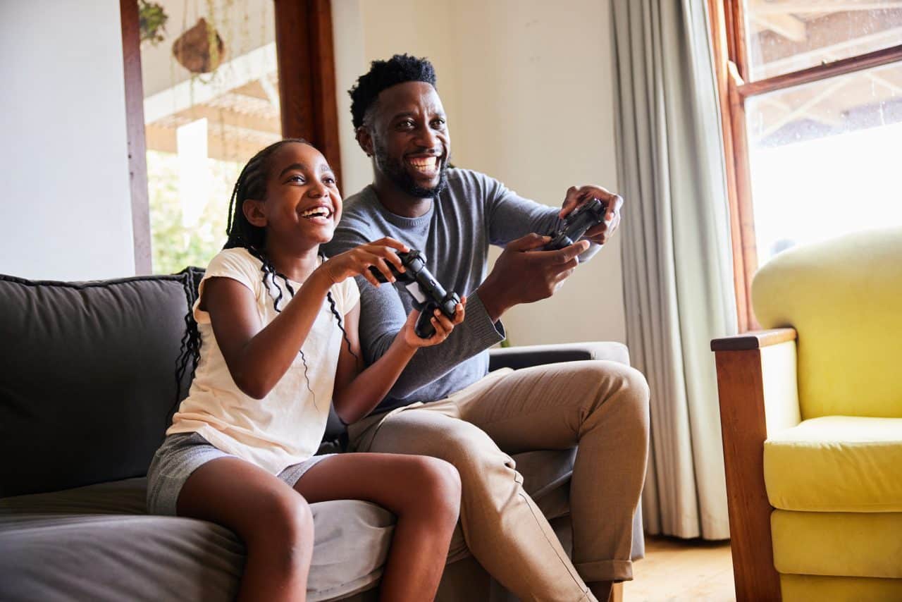 Little girl and her father laughing while playing video games together on a sofa in their living room at home
