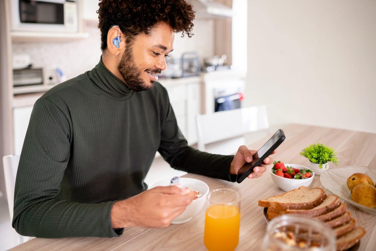 Young man with hearing aids looking at his smartphone over breakfast.